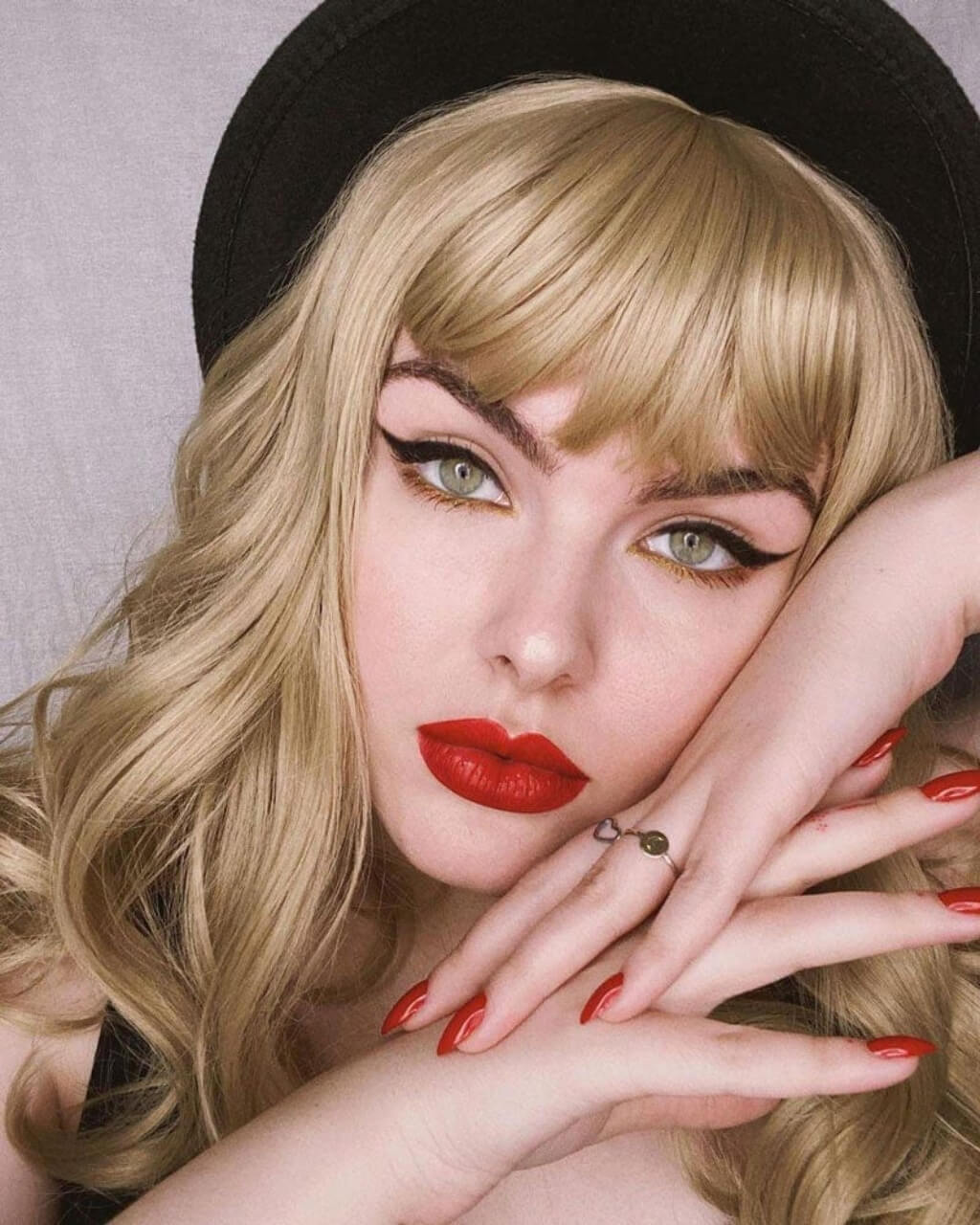 Woman with blonde hair, red lipstick, and a black hat posing with hands near her face. Ash blonde 50s style wig with short fringe and gentle waves: Trinity