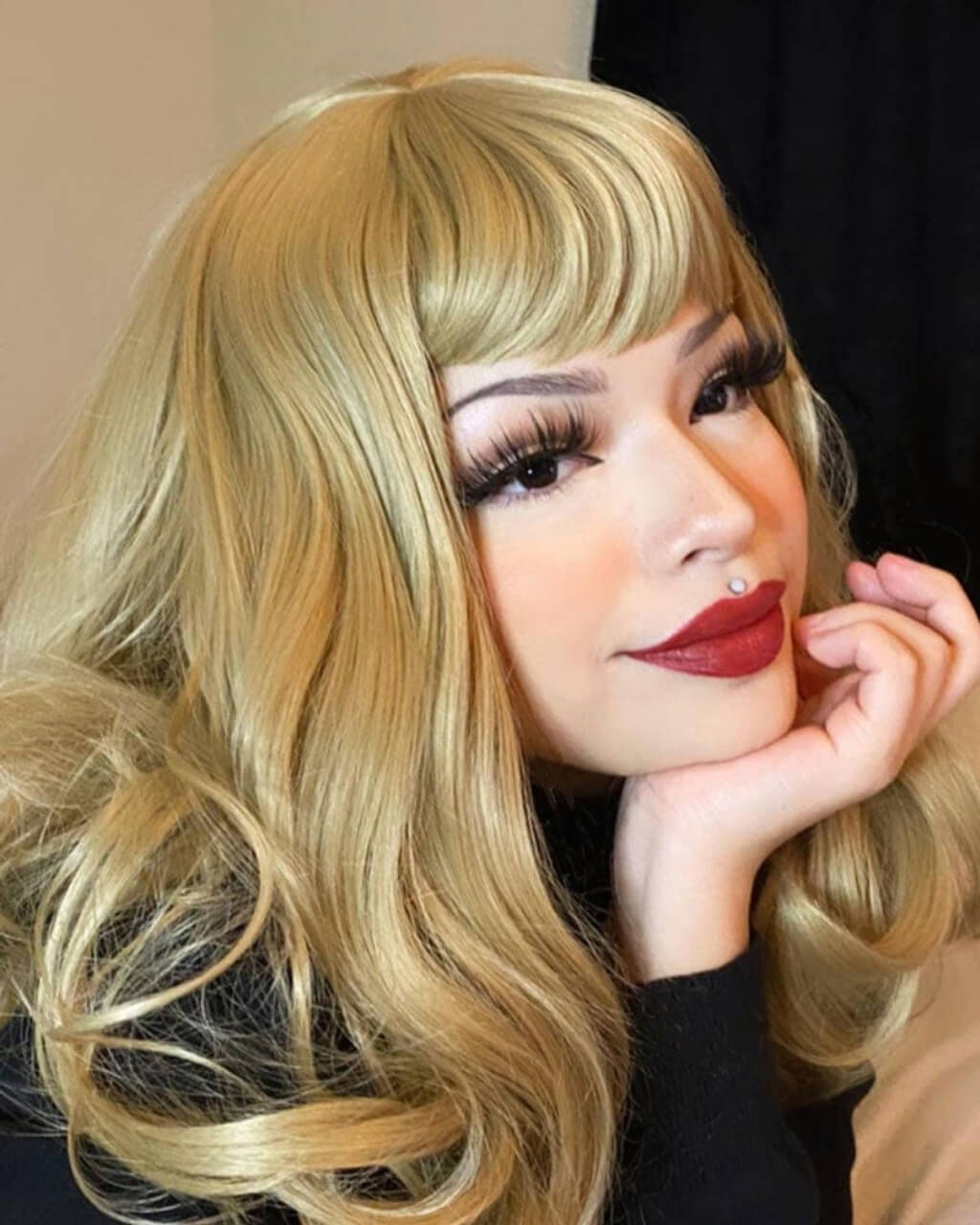 Person with blonde wig and red lipstick posing with hand on chin. Ash blonde 50s style wig with short fringe and gentle waves: Trinity