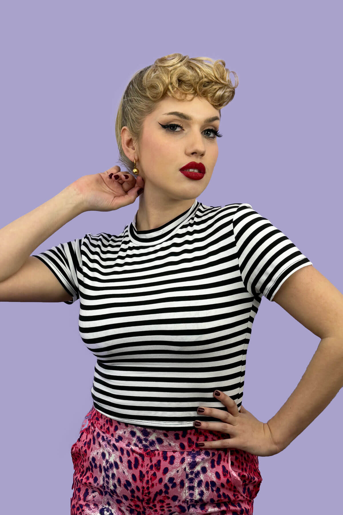 Woman wearing a black and white striped shirt with colorful pants against a purple background. Curly 1940s style clip-in fringe hairpiece/clip-in bangs: Bunny ash blonde 24 