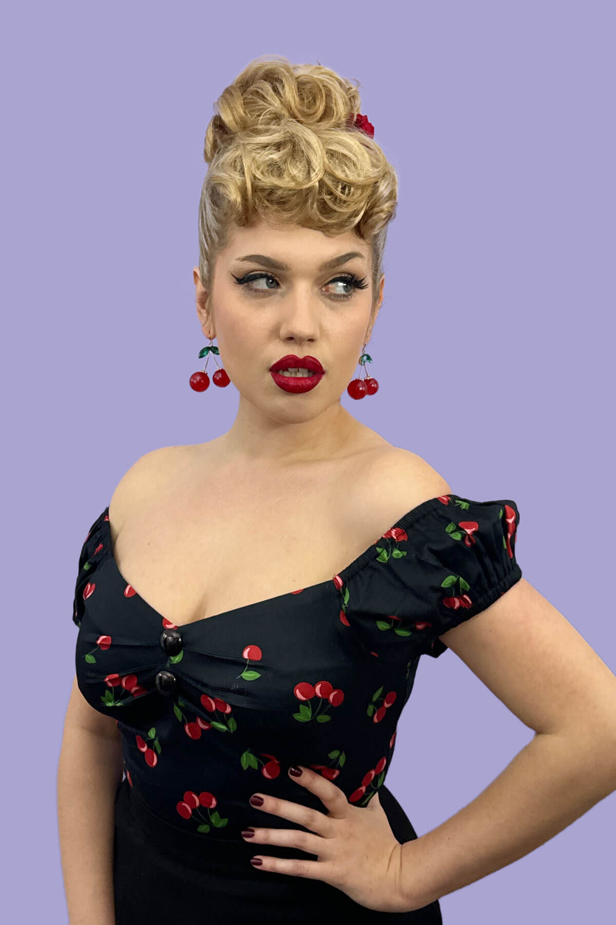 Woman wearing a black dress with red cherry patterns against a purple background. Curly bun hairpiece, rockabilly style: Toni black 1B