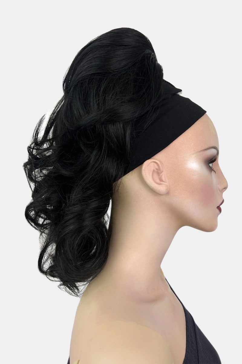 A woman wearing a curly ponytail extension hairpiece with loose, layered curls. She is seen from the shoulders up, with a backdrop featuring bokeh lights. Curly ponytail extension clip-on hairpiece: Tulisa black 1B