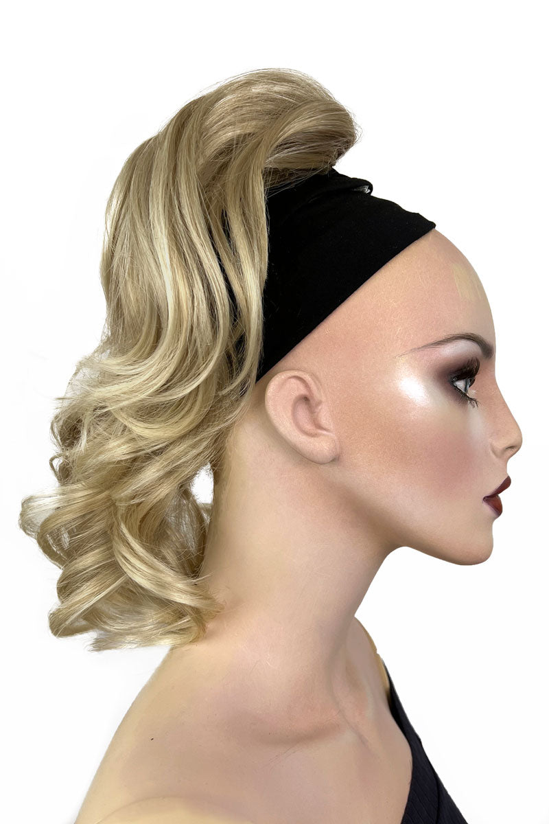 A woman wearing a curly ponytail extension hairpiece with loose, layered curls. She is seen from the shoulders up, with a backdrop featuring bokeh lights. Curly ponytail extension clip-on hairpiece: Tulisa blonde 613T24