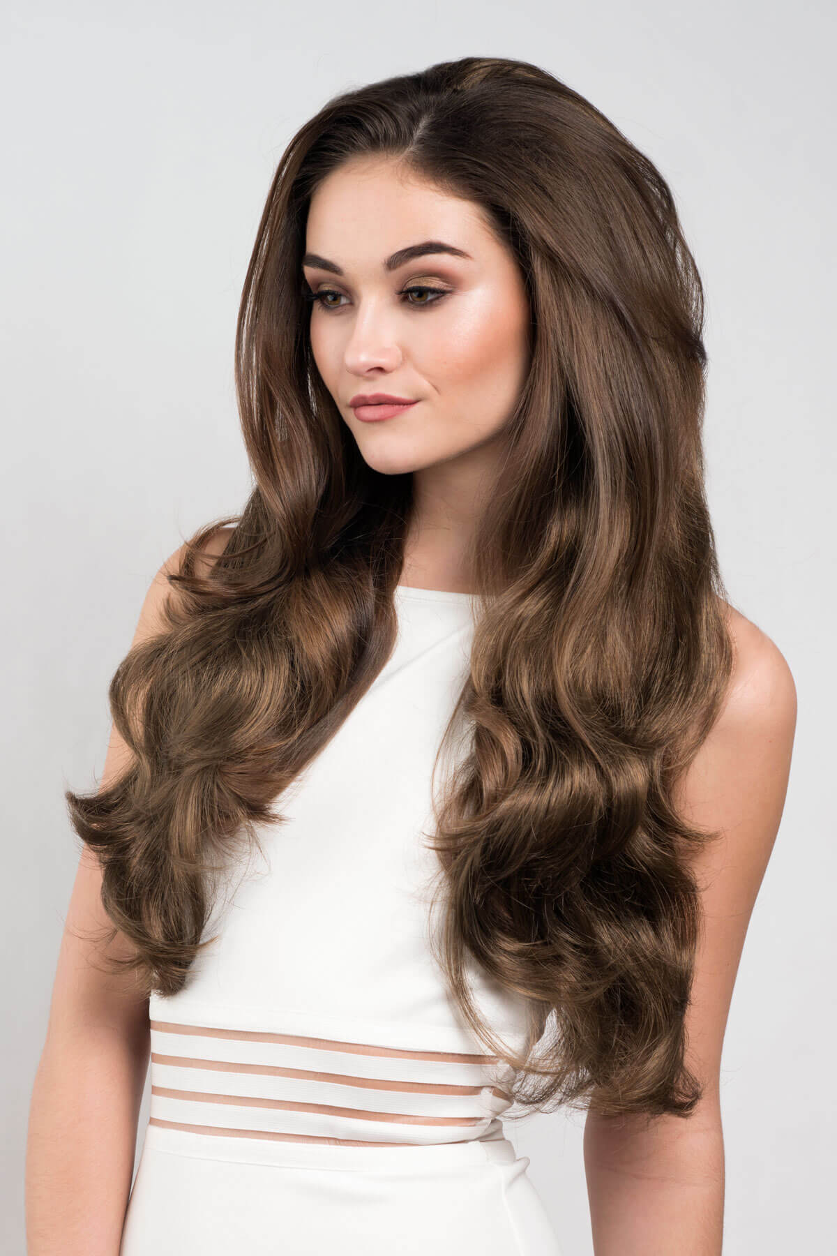 Woman with long, wavy brown hair wearing a white dress against a light gray background. Brown half wig hairpiece with long loose curls: Chelsea mocha brown 10