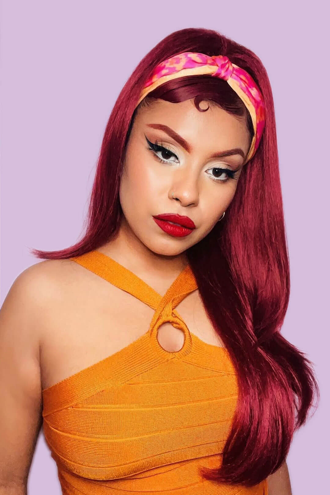 Woman with vibrant red hair and orange dress against a light purple background. 1960s style hair, high crown long straight and layered half wig: Juliette wine red 39