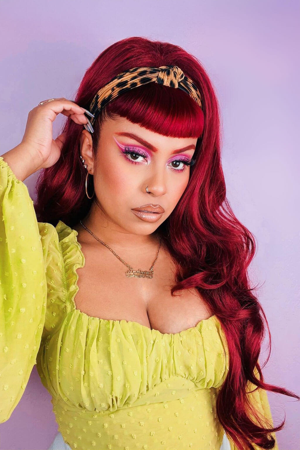 Woman with vibrant red hair and colorful makeup wearing a yellow top against a light purple background