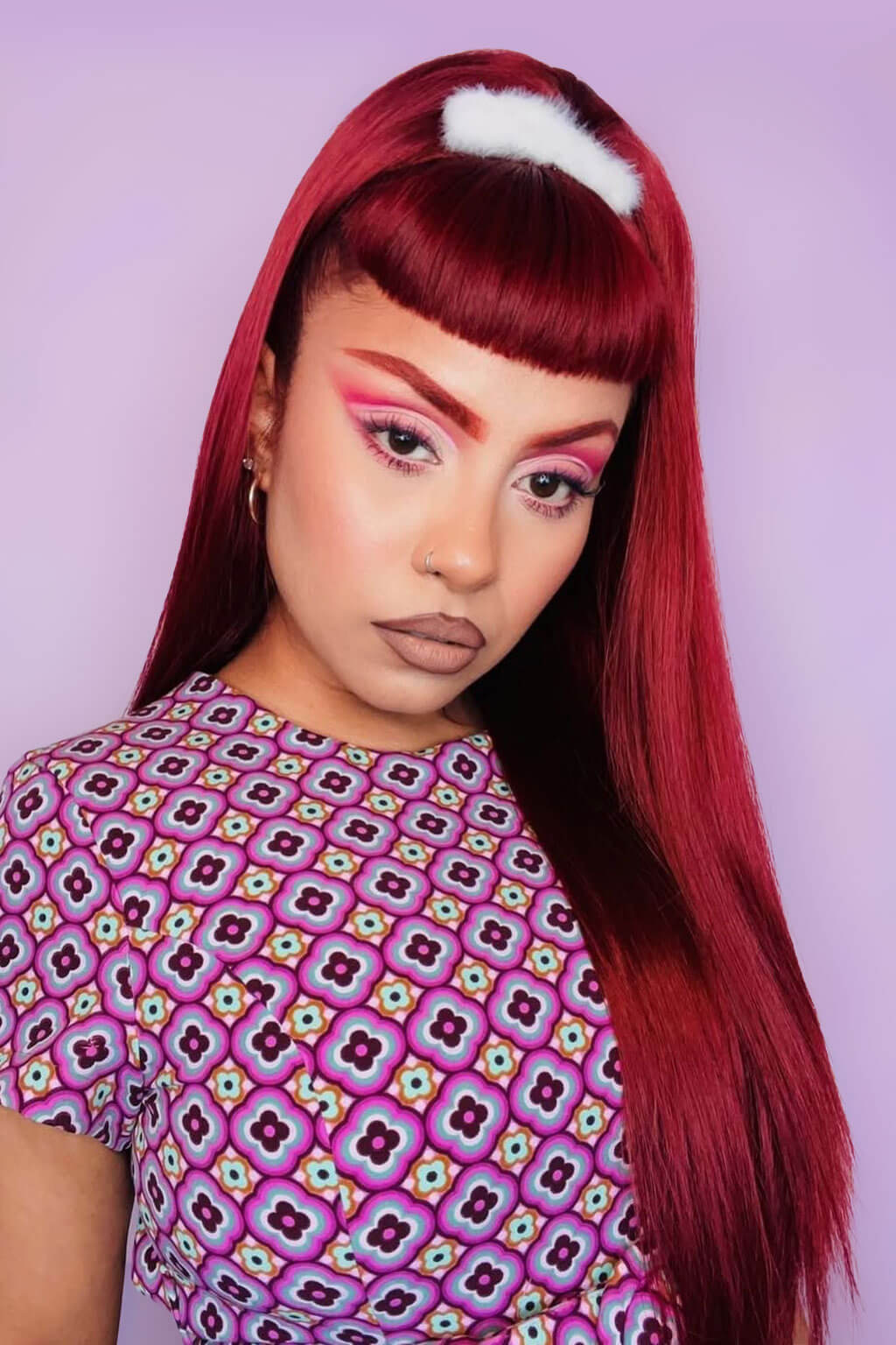 Person with vibrant red hair and colorful makeup wearing a patterned shirt against a light purple background. 1960s style hair, high crown long straight half wig: Andrea cherry red 39
