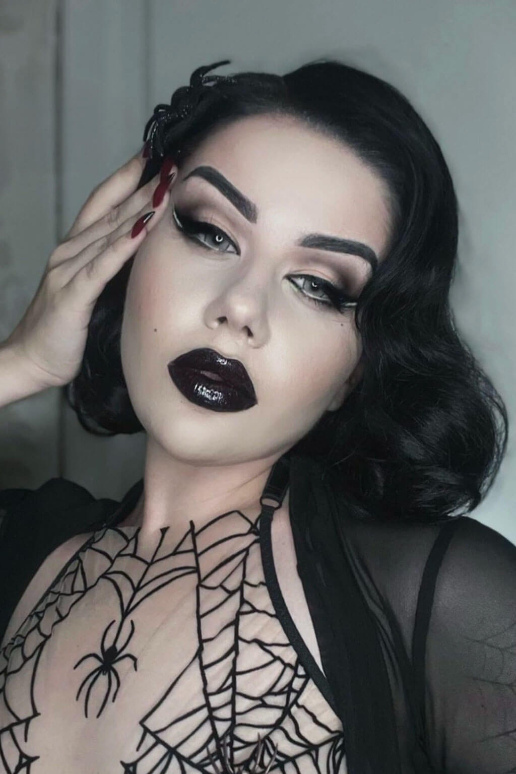 Woman with dark makeup and spider-themed sheer top against a neutral background. Black lacefront wig, pinup/vintage style, mid length with finger waves: Lilith