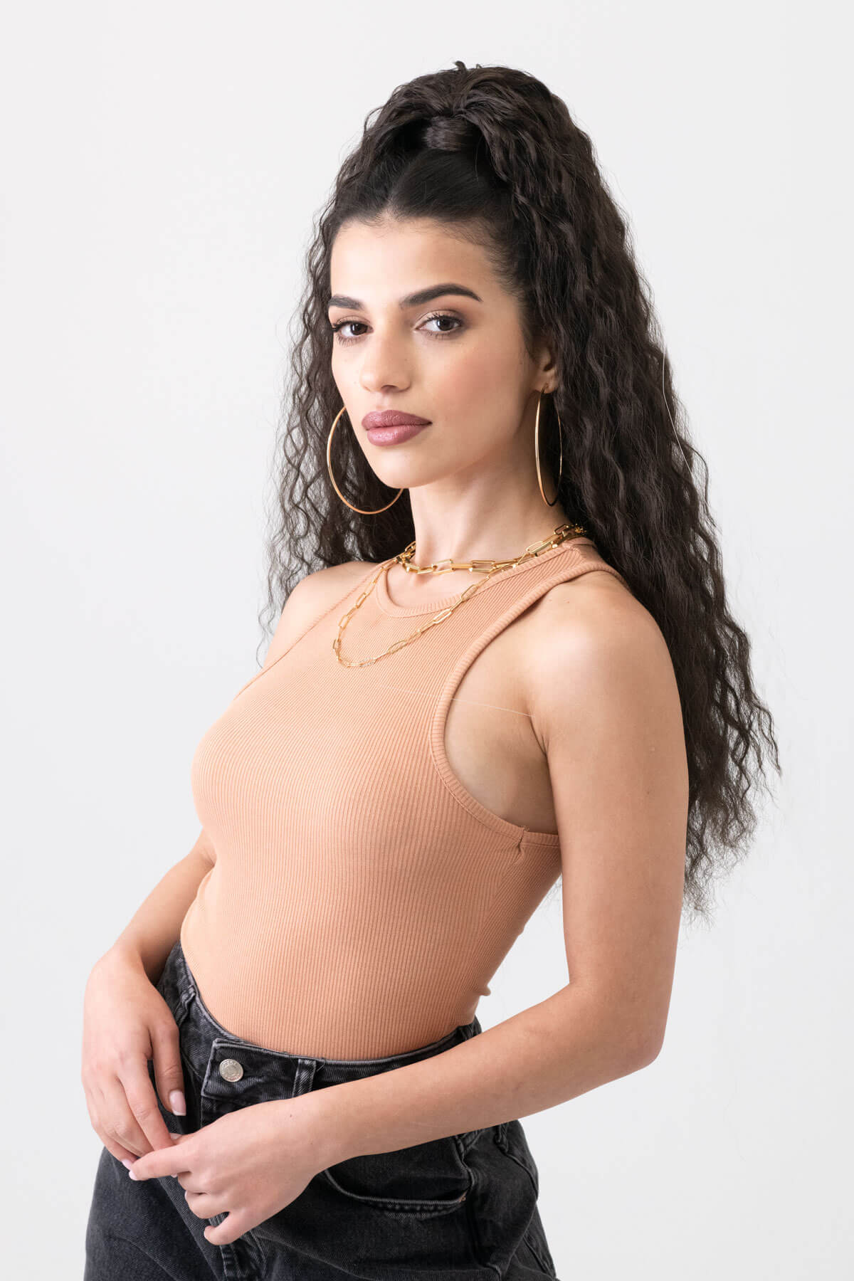 Woman wearing a beige tank top and black pants on a white background.  Extra long ponytail extension with long loose spirals: Kaia dark brown 4