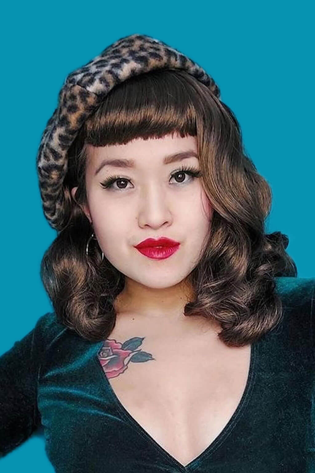 Woman wearing a leopard print hat and green velvet top against a blue background. Brown pinup style wig, curled with short fringe: Carolina mocha brown 10