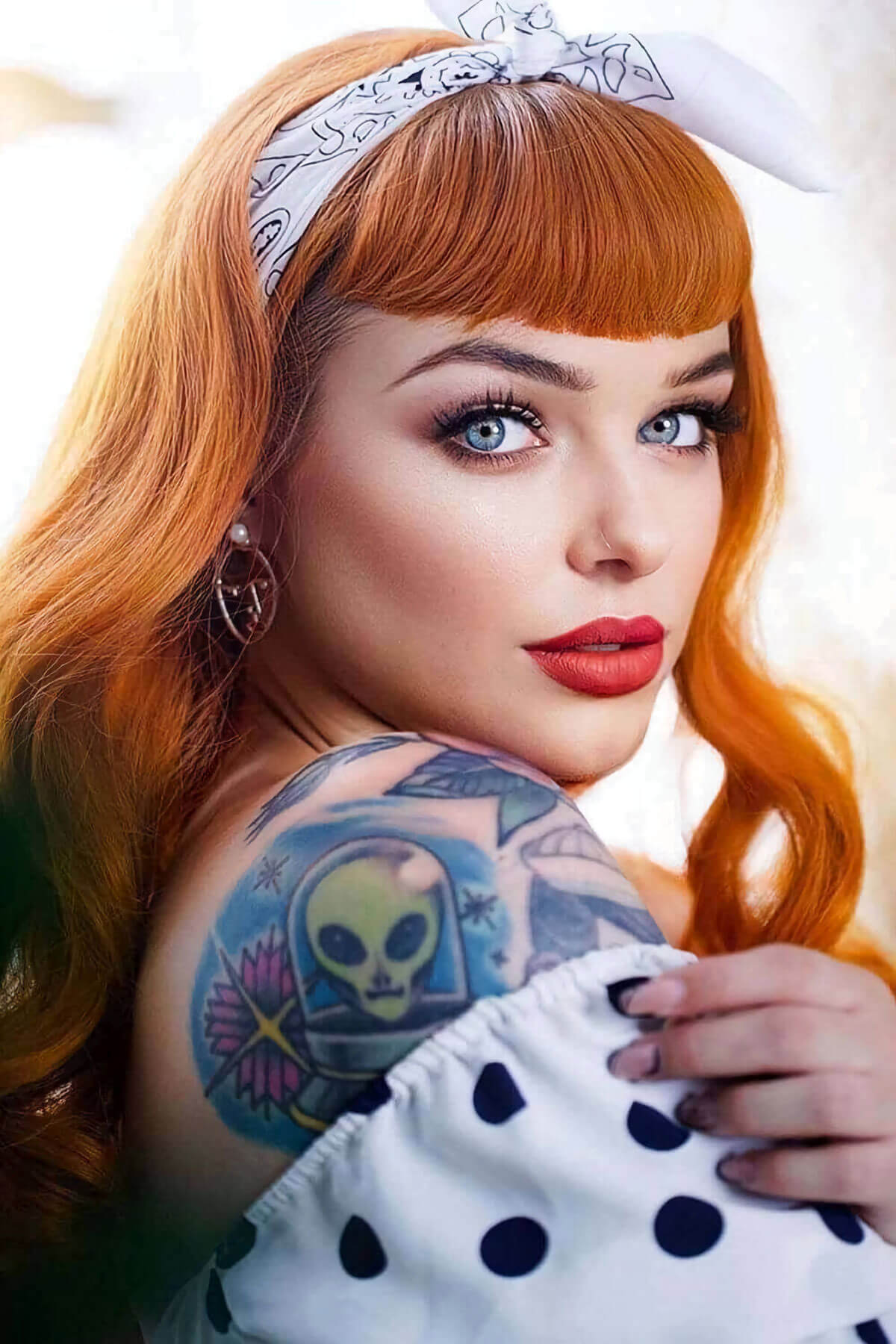 Woman with red hair and tattoos on her shoulder wearing a white polka dot dress. Ginger wig, pinup style with short fringe and gentle waves, 1950s style: Vida