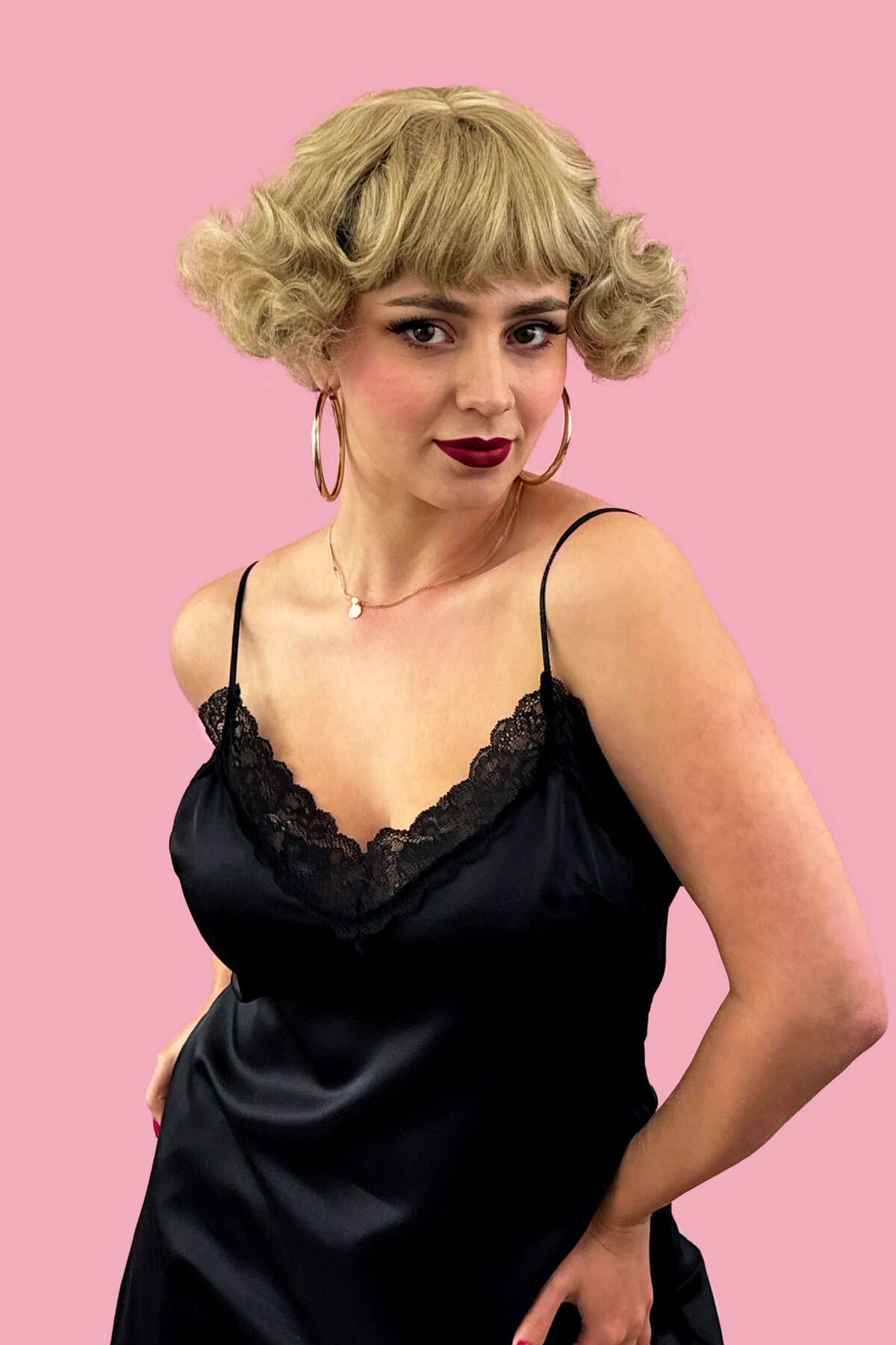 Woman wearing a black satin slip dress with lace detailing against a pink background. A short blonde vintage style wig with waves: Joy