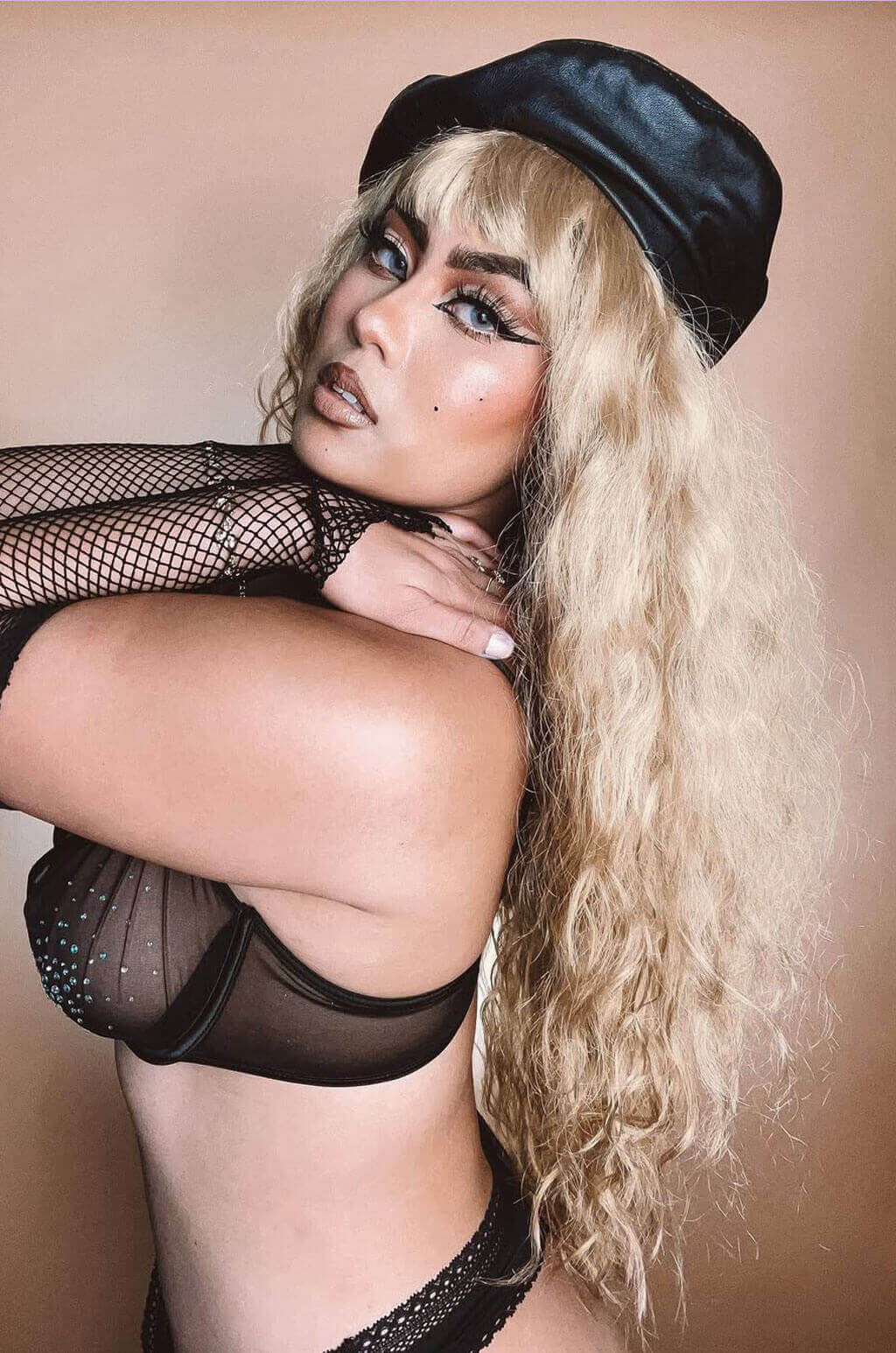 Woman wearing a black cap and fishnet stockings against a beige background. Woman wearing a black cap and fishnet top against a beige background. Long wig with crimped mermaid waves: Astrid