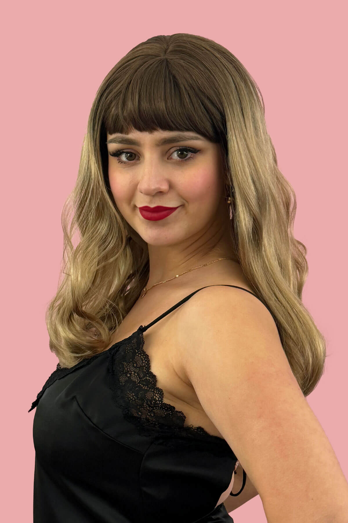 Woman with long wavy hair wearing a black dress against a pink background. Long brown wavy bob (LOB) wig blended brown to blonde: Avery