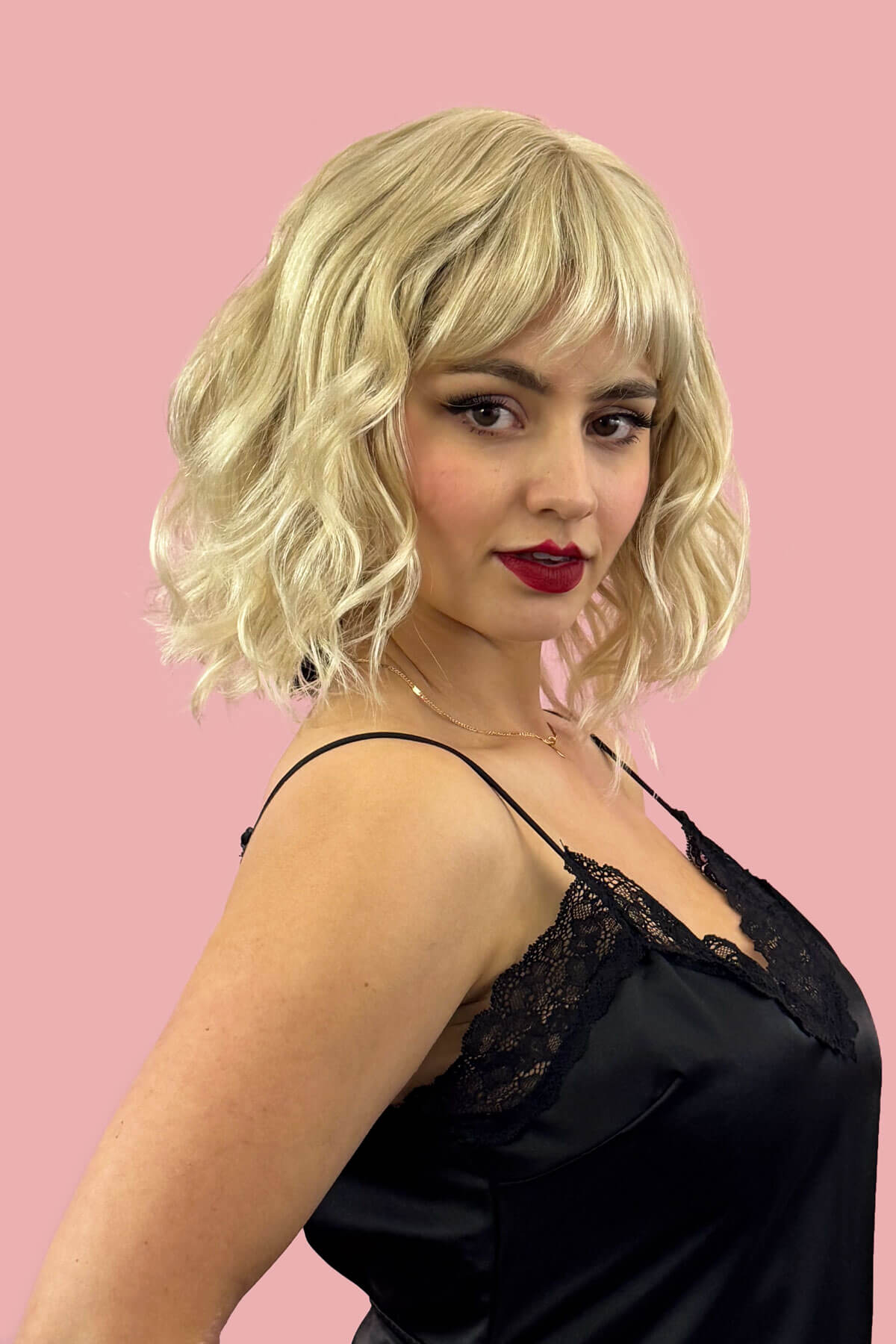 Woman with blonde wig and red lipstick wearing a black lace top against a pink background. Blonde long wavy bob (lob) wig, ash blonde blending to light blonde: Pearl blonde 613T24