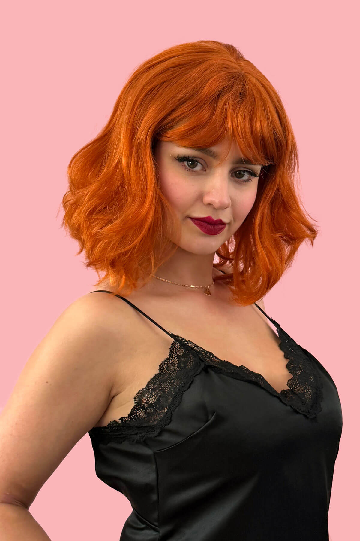 Woman with red hair wearing a black lace top against a pink background. Ginger long wavy bob (lob) wig with swept fringe: Rusty