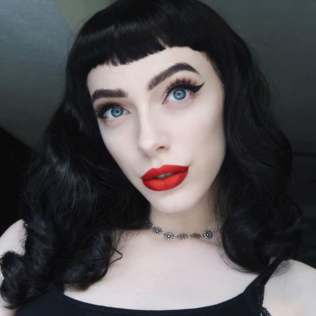 Person with black hair and red lipstick wearing a black top against a dark background. Black pin-up style wig, old Hollywood waves with short fringe: Bettie
