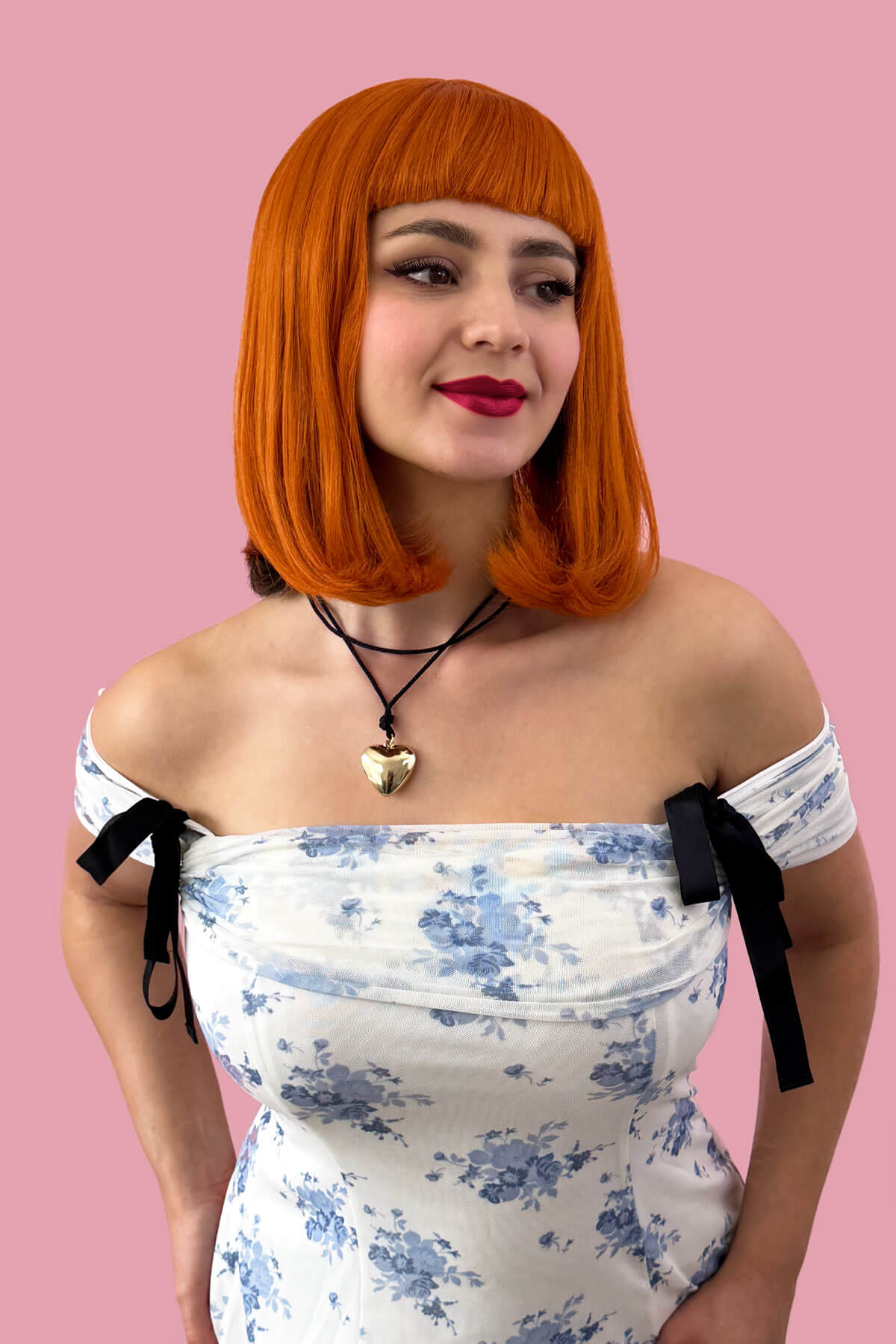 Woman with orange wig wearing a floral off-shoulder dress against a pink background. Ginger long bob wig with a short retro style fringe: Bonnie