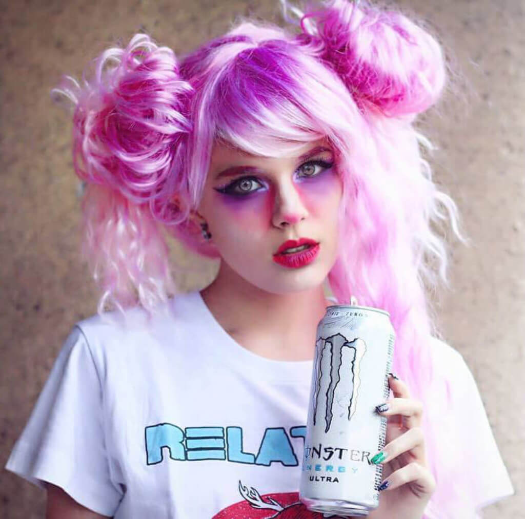 A person with bright pink hair styled in two buns holds a can. They are wearing a white graphic t-shirt against a textured neutral background. Crimped pink wig, long, in two colour pink ombre: Peony