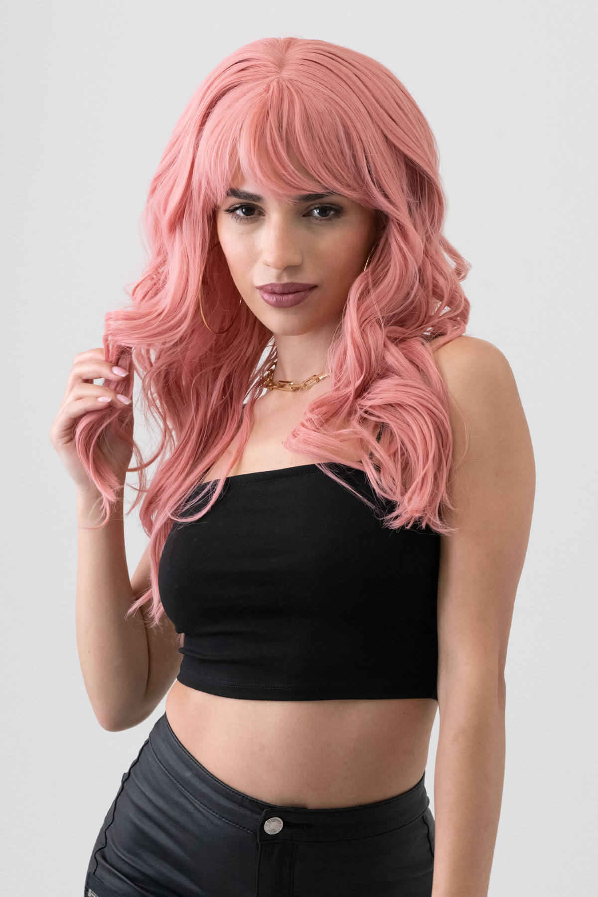 Woman with pink wig wearing a black top and black pants on a white background. Dusty pastel pink wig with long cascading waves: Rosalie