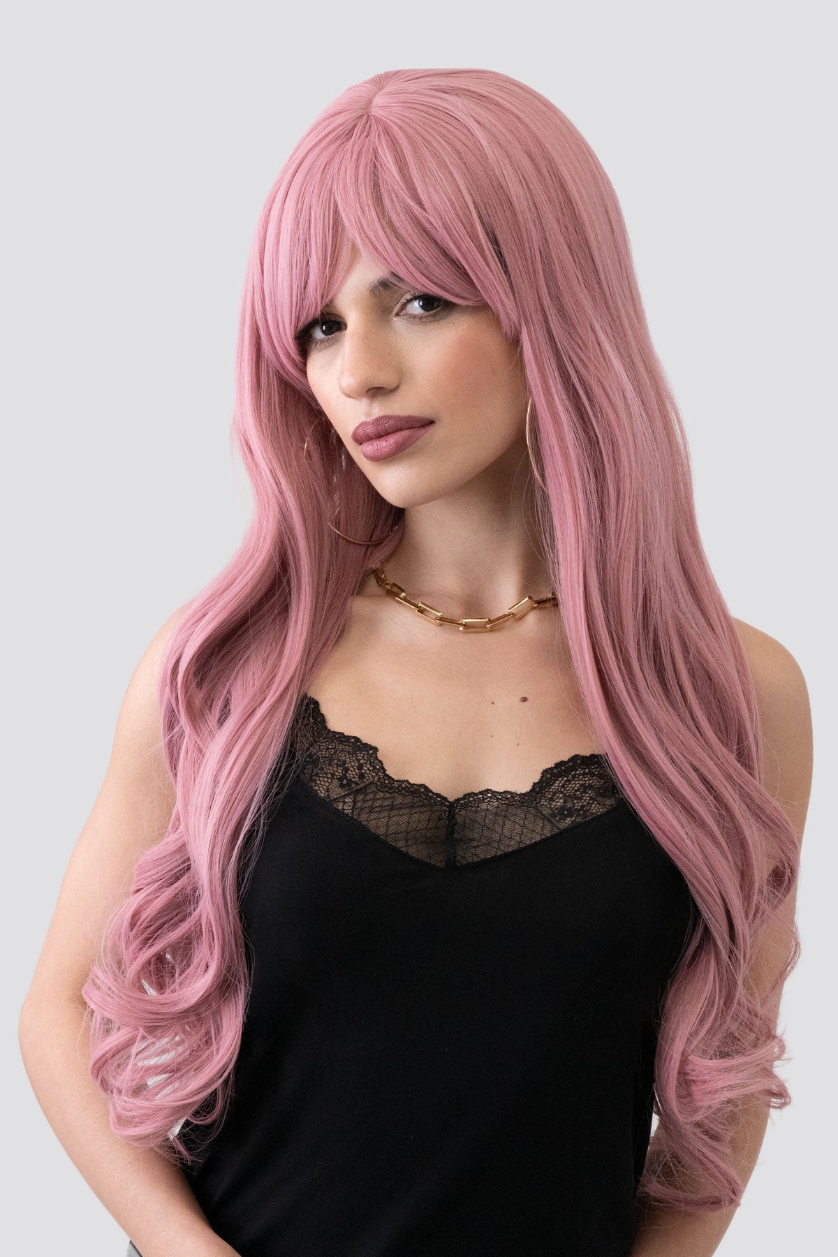 Woman with long pink wig wearing a black top on a light gray background. Long wavy pink wig with sweeping fringe: Millicent