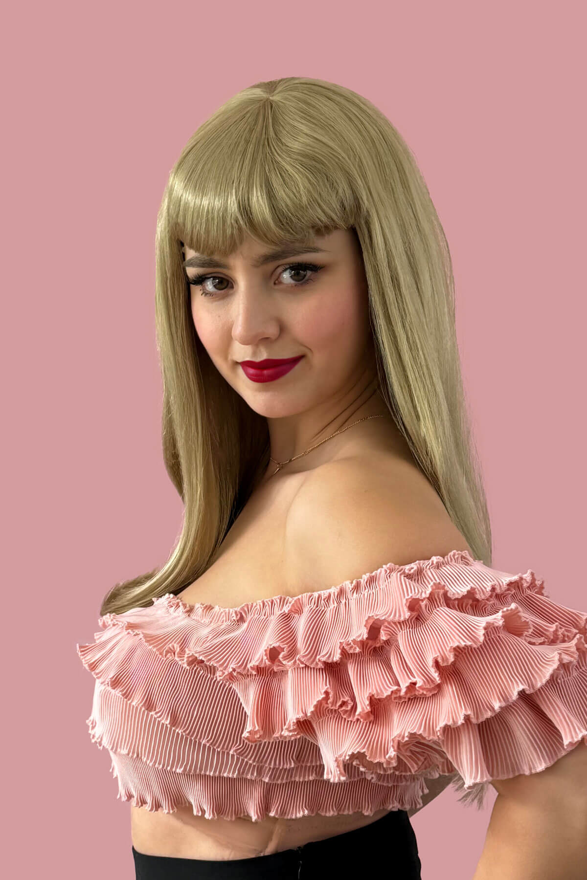 Woman wearing a pink off-shoulder top with ruffles against a pink background. Long blonde wig with straight hair and a short, straight fringe: Nicole ash blonde 24