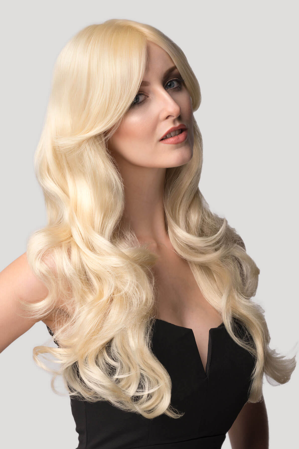 Woman with long blonde wig on a plain background. Blonde wig with big loose curls and sweeping fringe, extra long: Carly champagne blonde 613