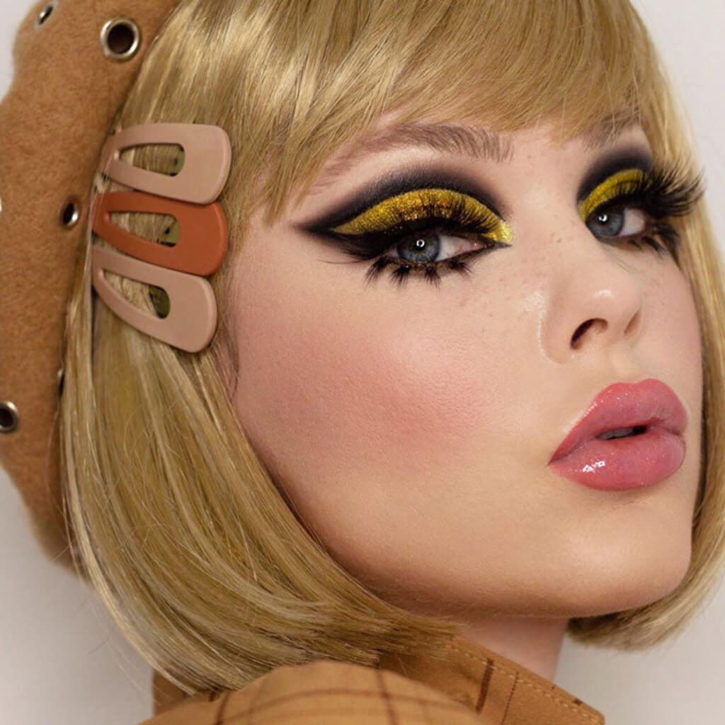 Close-up of a person with bold makeup and a brown headband with circular elements. Blunt blonde bob wig with fringe: Gracie 24H613