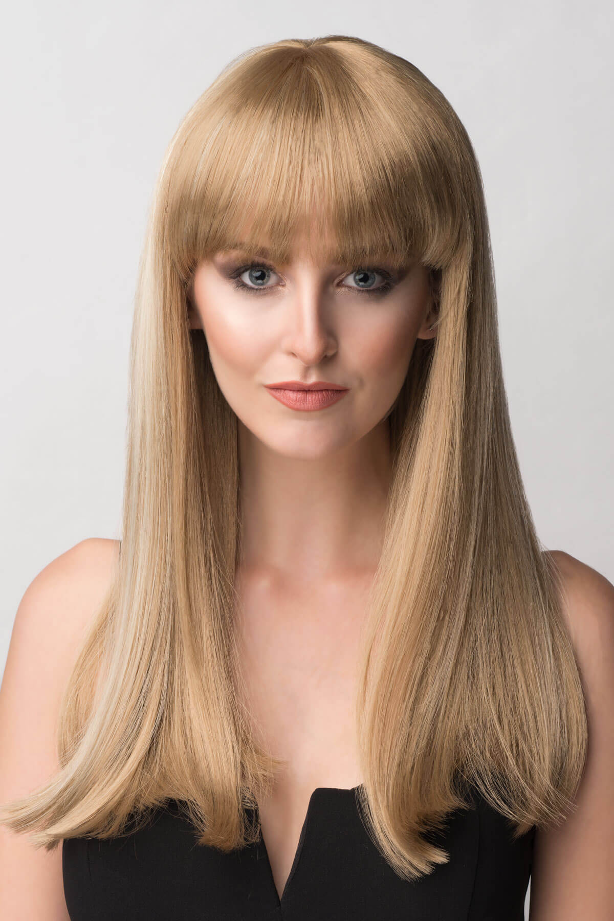 Woman with long blonde hair and bangs against a plain background. Long blonde wig with Highlights and straight fringe: Tilly