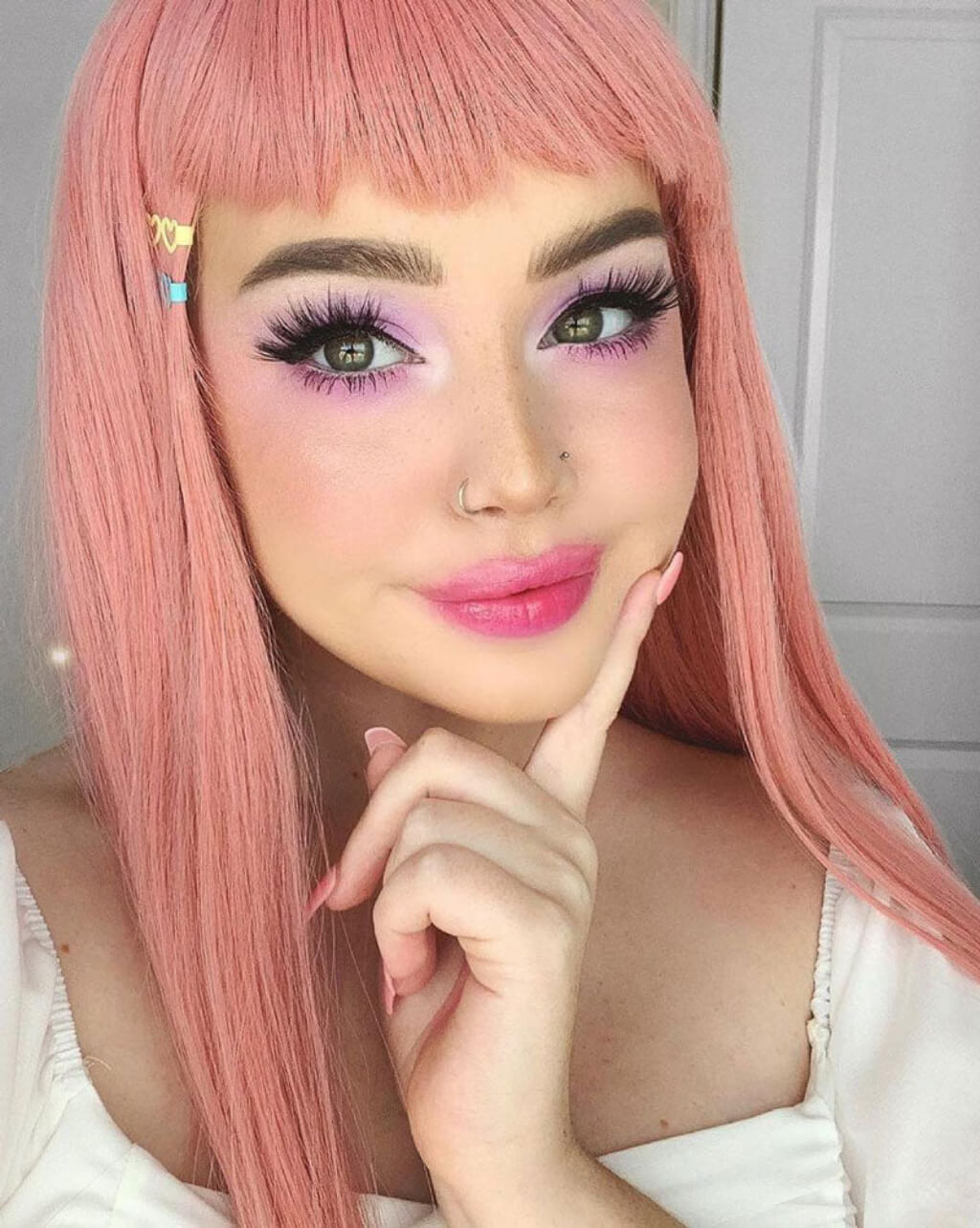 Person with pink hair and colourful makeup posing with hand on chin. Long Pink Wig With Short, Straight Fringe: Stephanie