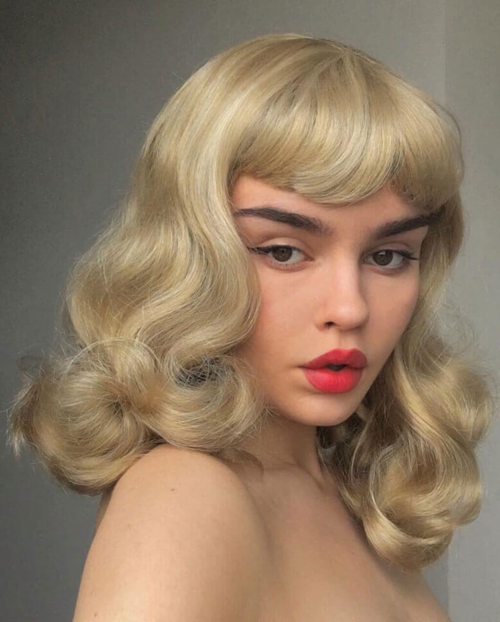 Woman with a blonde wig styled in vintage curls against a plain background. Blonde pinup wig, curled with short fringe, 1950s style: Cora blonde 24H613