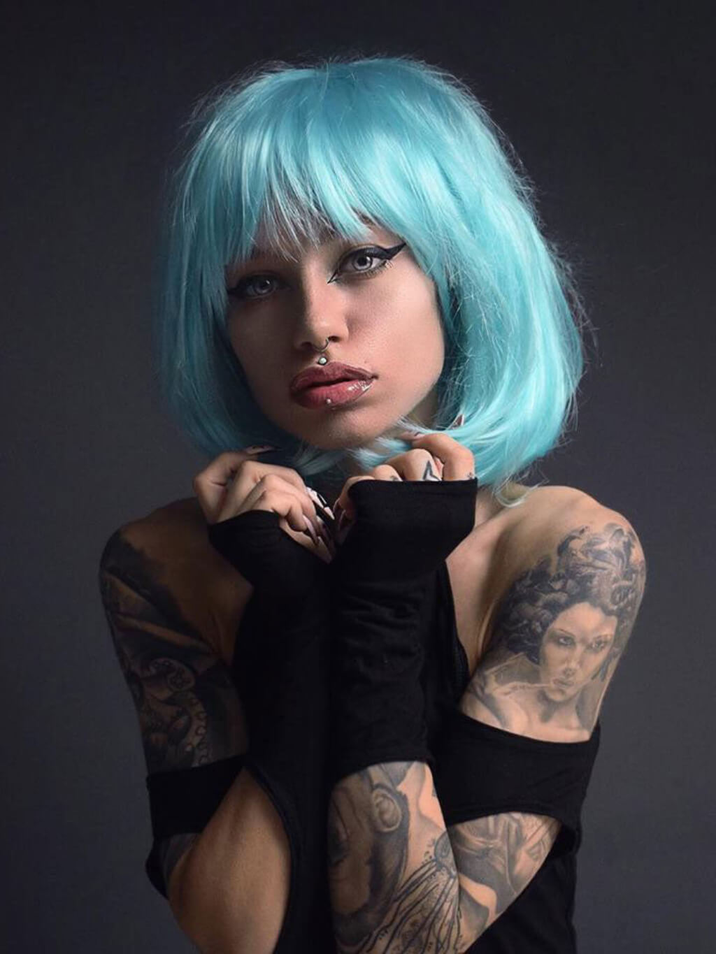 Person with blue hair and tattoos wearing a black outfit against a dark background. Turquoise bob wig  with full fringe: Iris
